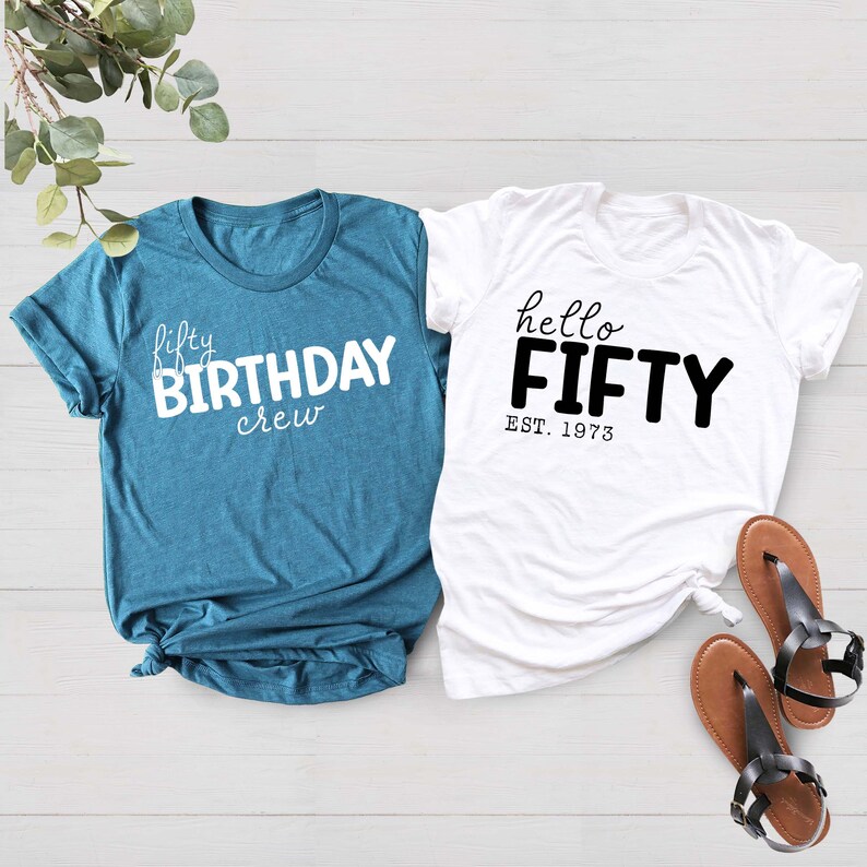 May include: Two t-shirts, one teal blue and one white, with birthday greetings. The teal blue shirt says "fifty birthday crew" in white text. The white shirt says "hello fifty est. 1973" in black text.