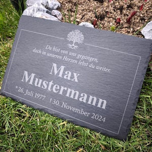 Könnte beinhalten: Rechteckige, dunkelgraue Gedenktafel aus Schiefer mit weiß graviertem Text. Der Text enthält einen Namen, Daten und eine kurze Nachricht. Die Tafel liegt auf grünem Gras mit Felsen im Hintergrund.