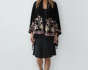 May include: A black velvet kimono-style jacket with floral embroidery in pink, beige, and green. The jacket has a shawl collar and wide sleeves. The model is wearing a black dress and white sneakers.