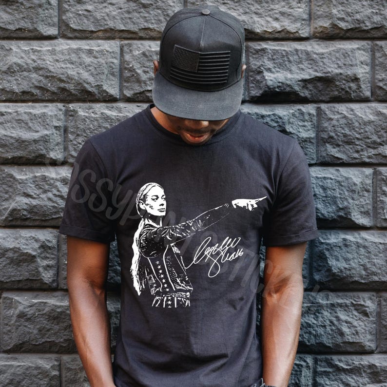 May include: A black short-sleeved t-shirt featuring a white graphic print of a person with a ponytail pointing to the right, accompanied by a signature. The shirt is paired with a black baseball cap that has a black American flag patch on the front.