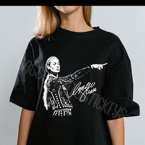 May include: A black short-sleeved t-shirt featuring a white graphic print of a person with long hair pointing to the side. The design includes a stylized signature script below the figure. The shirt has a crew neckline and a relaxed fit.