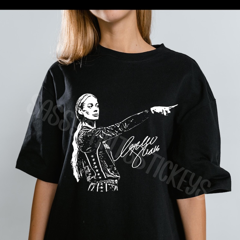 May include: A black short-sleeved t-shirt featuring a white graphic print of a person with long hair pointing to the side. The design includes a stylized signature script below the figure. The shirt has a crew neckline and a relaxed fit.