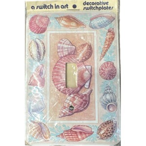 Puede incluir: Placa de interruptor decorativa con diseño de conchas marinas. La placa tiene una abertura central rectangular y está rodeada de varias ilustraciones de conchas en tonos rosa, beige y azul. El texto "a switch in art" y "decorative switchplates" es visible.