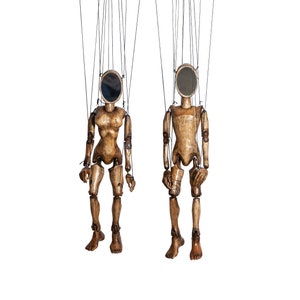 May include: Two wooden marionette puppets with oval mirrors for faces hang from a black control bar. The puppets have articulated limbs and are connected by strings. The puppets are made of light brown wood.