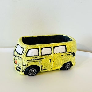 May include: A yellow ceramic planter shaped like a vintage Volkswagen bus. The planter has black painted details and white windows.