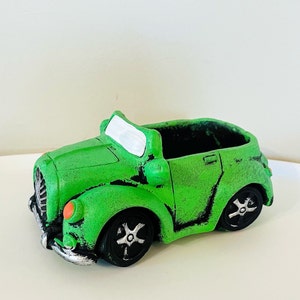 May include: A green vintage car-shaped planter with black wheels and silver accents. The planter has an open top for planting and orange headlights. The car has a distressed finish, adding to its vintage charm.