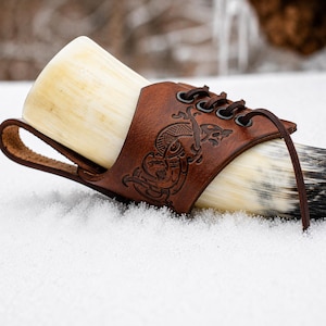 May include: A Viking drinking horn made of natural horn, held in place by a brown leather holder. The leather holder is embossed with a detailed design and laced with a brown cord. The horn rests on a bed of snow.