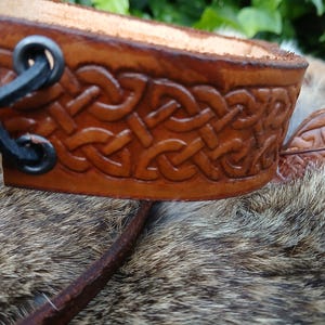 May include: Close-up of a brown leather cuff with a Celtic knot design. The cuff is intricately carved and features a dark brown border. Black metal rings and leather straps are visible on the side. The background is blurred.