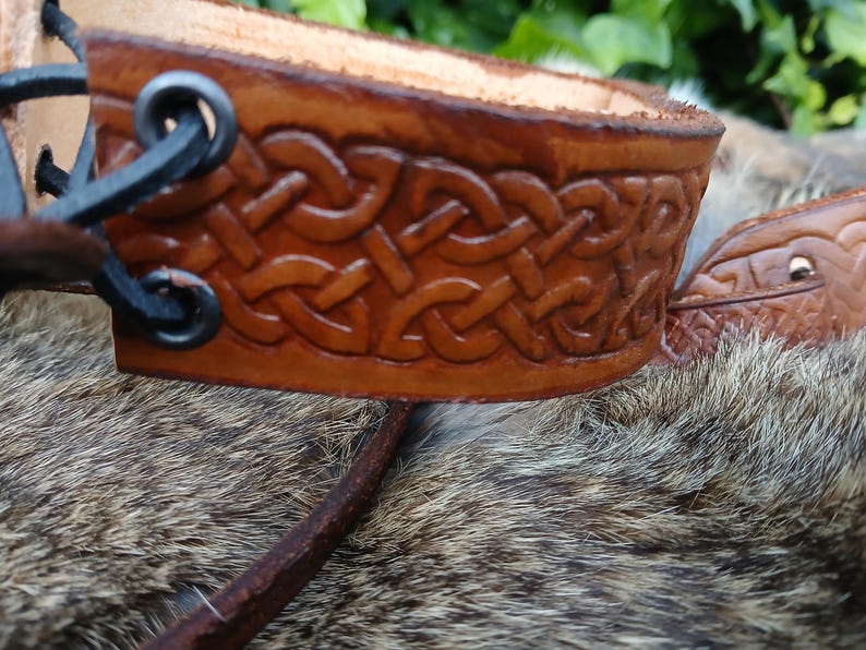 May include: Close-up of a brown leather cuff with a Celtic knot design. The cuff is intricately carved and features a dark brown border. Black metal rings and leather straps are visible on the side. The background is blurred.
