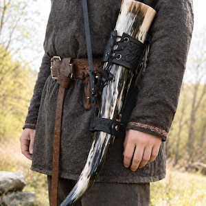 May include: A Viking-style drinking horn with a black leather holder and strap. The horn is black and white, and the holder is laced up. The person is wearing a brown tunic and pants.