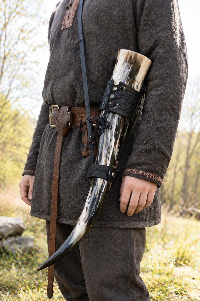 May include: A Viking-style drinking horn with a black leather holder and strap. The horn is black and white, and the holder is laced up. The person is wearing a brown tunic and pants.