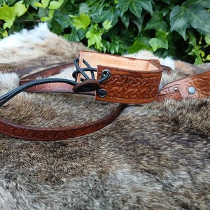May include: Hand-tooled leather wrist cuff and belt in shades of brown. The cuff features a Celtic knot design and black lacing. The belt has a matching embossed pattern. The items are displayed on a fur-like surface.