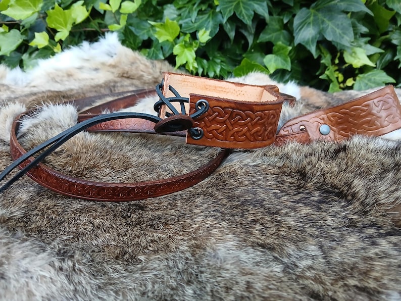 May include: Hand-tooled leather wrist cuff and belt in shades of brown. The cuff features a Celtic knot design and black lacing. The belt has a matching embossed pattern. The items are displayed on a fur-like surface.