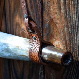 May include: A brown leather horn holder with Celtic knotwork design, secured with rivets. The holder is attached to a polished horn with a dark brown tip. The background is a weathered wooden surface.