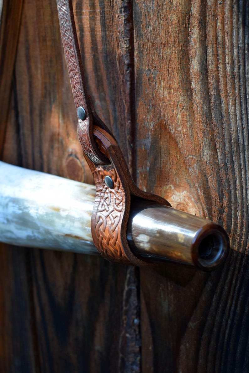 May include: A brown leather horn holder with Celtic knotwork design, secured with rivets. The holder is attached to a polished horn with a dark brown tip. The background is a weathered wooden surface.