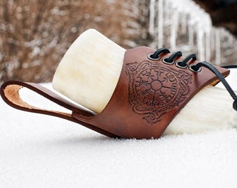 May include: A Viking drinking horn with a brown leather holder. The holder features a detailed embossed design and black lacing. The horn is a light cream color and rests on a snowy surface.