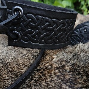 May include: Close-up of a black leather wrist cuff with a Celtic knot design. The cuff is made of thick leather and has a textured surface. The background is blurred, with green foliage and fur.