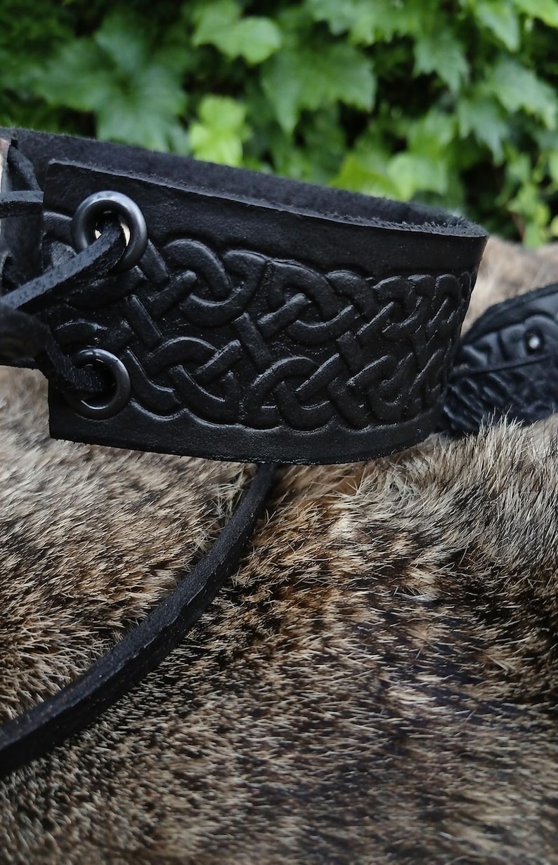 May include: Close-up of a black leather wrist cuff with a Celtic knot design. The cuff is made of thick leather and has a textured surface. The background is blurred, with green foliage and fur.