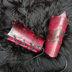 May include: Pair of burgundy leather arm guards with decorative edging and metal rivets. The arm guards are laced together with black leather cord. The leather has a distressed appearance.