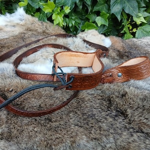May include: A brown leather stirrup with a matching strap, featuring embossed Celtic knot designs. The stirrup is connected with black leather ties. The items are displayed on a fur-like surface, with green foliage in the background.