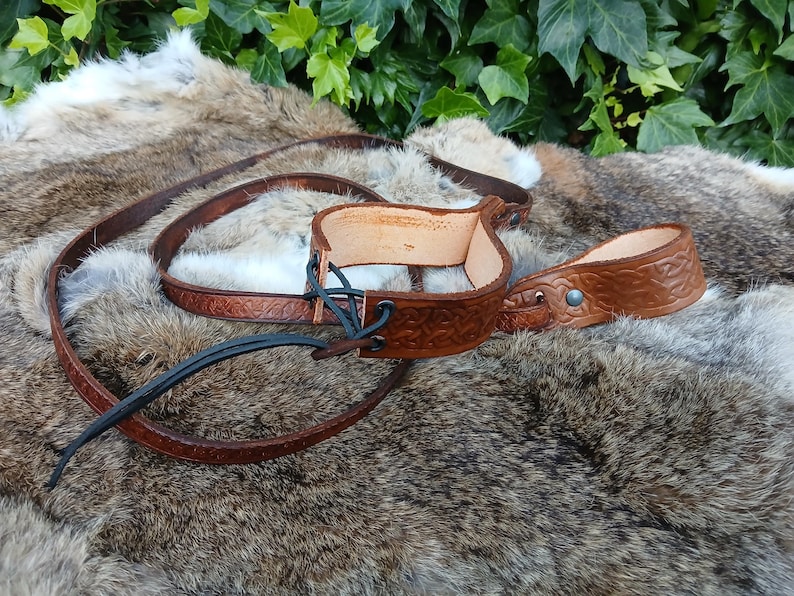 May include: A brown leather stirrup with a matching strap, featuring embossed Celtic knot designs. The stirrup is connected with black leather ties. The items are displayed on a fur-like surface, with green foliage in the background.