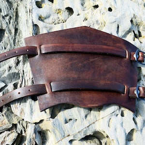 Peut inclure: Une protection d'avant-bras en cuir marron avec deux sangles et des boucles. La protection est conçue pour protéger l'avant-bras des blessures.