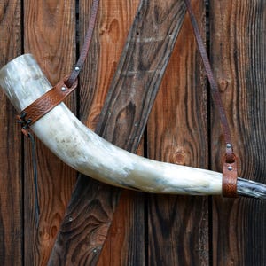 May include: A large, curved drinking horn made of polished horn, with a brown leather strap and decorative band. The horn is suspended against a rustic wooden background, showcasing its natural color variations and texture.