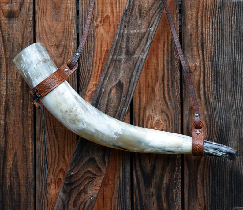 May include: A large, curved drinking horn made of polished horn, with a brown leather strap and decorative band. The horn is suspended against a rustic wooden background, showcasing its natural color variations and texture.