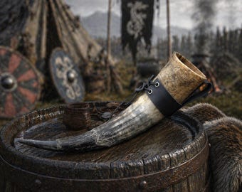 May include: A Viking-style drinking horn with a black leather strap and eyelets, resting on a wooden barrel. The horn is light brown and tan, with a dark tip. A small, dark brown cup sits nearby.