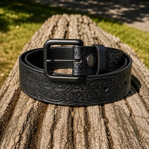May include: A black leather belt with a dark metal buckle. The belt has an embossed Celtic knot pattern and a matching loop with a decorative metal stud. The belt is coiled on a weathered wooden surface.