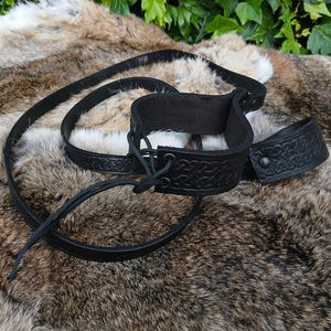 May include: Black leather wrist and ankle restraints with embossed Celtic knot designs. The set includes two cuffs and a connecting strap. The cuffs have a buckle closure and are shown on a fur-like surface.