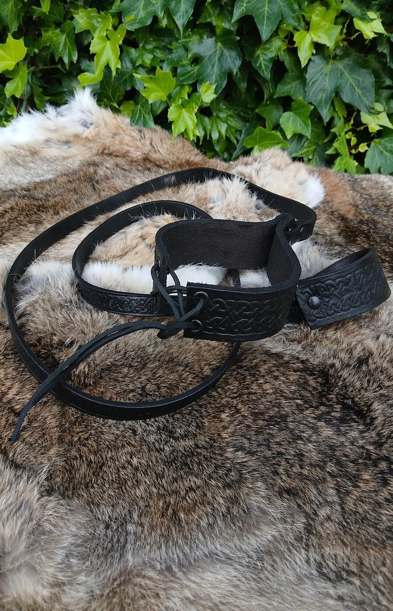 May include: Black leather wrist and ankle restraints with embossed Celtic knot designs. The set includes two cuffs and a connecting strap. The cuffs have a buckle closure and are shown on a fur-like surface.