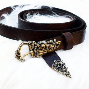 Peut inclure: Une ceinture en cuir marron foncé avec une boucle décorative dorée ornée d'un motif de nœud celtique complexe. La ceinture est enroulée, mettant en valeur sa texture et la boucle détaillée. Un accessoire élégant.