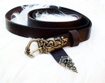 May include: A dark brown leather belt with a decorative gold-colored buckle featuring an intricate Celtic knot design. The belt is coiled, showcasing its texture and the detailed buckle. A stylish accessory.