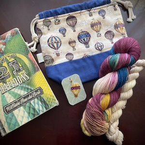 May include: An assortment of craft supplies featuring a book titled "21 Balloons", a drawstring bag with hot air balloon pattern, a skein of colorful yarn, and a hot air balloon charm. The book cover has a vintage design with colorful balloons.