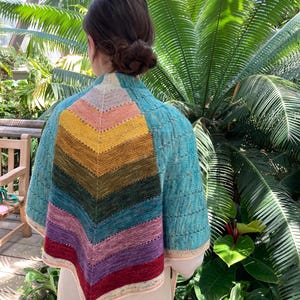 May include: A colorful knitted shawl with a chevron pattern in shades of yellow, orange, pink, green, blue, purple, and red. The shawl has a turquoise body and a tassel at the bottom. The garment is worn over a cream-colored top.
