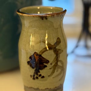 May include: A small, handcrafted ceramic vase with a light green glaze. The vase features a dark brown floral design and abstract brushstrokes. The rim and base have a darker, contrasting color. The vase is approximately 4 inches tall.