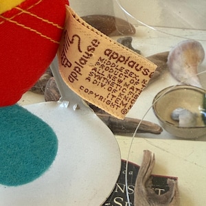 May include: A plush toy featuring a red and yellow fabric body with a teal circular patch. A beige fabric tag is attached, printed with the text 'applause', 'MIDDLESEX, N.J.', 'PRODUCT OF KOREA', 'ALL NEW MATERIALS', 'SYNTHETIC FIBERS', and 'A DIV. OF KNICKERBOCKER'.