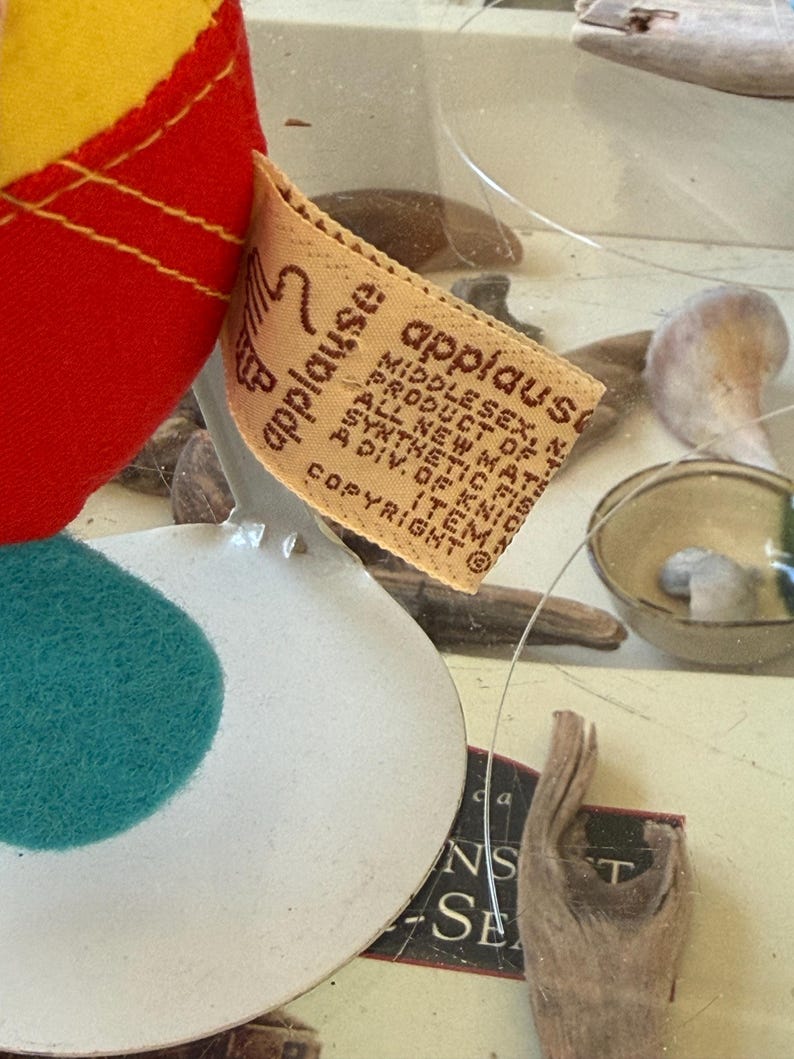 May include: A plush toy featuring a red and yellow fabric body with a teal circular patch. A beige fabric tag is attached, printed with the text 'applause', 'MIDDLESEX, N.J.', 'PRODUCT OF KOREA', 'ALL NEW MATERIALS', 'SYNTHETIC FIBERS', and 'A DIV. OF KNICKERBOCKER'.