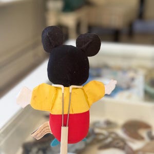 May include: A plush character puppet featuring a black head with round ears, a yellow shirt, and red pants. The puppet is mounted on a white metal stand with a Y-shaped support frame that holds the figure upright. A small fabric tag is attached to the side.