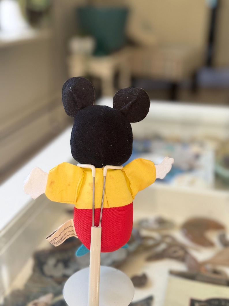 May include: A plush character puppet featuring a black head with round ears, a yellow shirt, and red pants. The puppet is mounted on a white metal stand with a Y-shaped support frame that holds the figure upright. A small fabric tag is attached to the side.