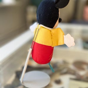 May include: A plush Mickey Mouse figure with a black head, yellow shirt, and red shorts, mounted on a white metal display stand. The figure has white felt hands and is positioned in profile, showing the back of the head and the side of the body.