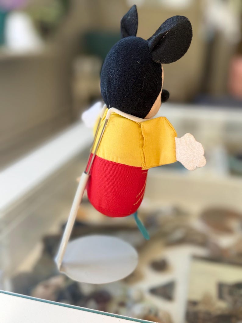 May include: A plush Mickey Mouse figure with a black head, yellow shirt, and red shorts, mounted on a white metal display stand. The figure has white felt hands and is positioned in profile, showing the back of the head and the side of the body.