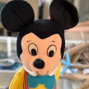 May include: A plush Mickey Mouse doll featuring large black ears, a peach-colored face, and a black nose. The doll wears a yellow shirt, a bright blue bowtie, and red pants held up by multicolored striped suspenders with a white collar.