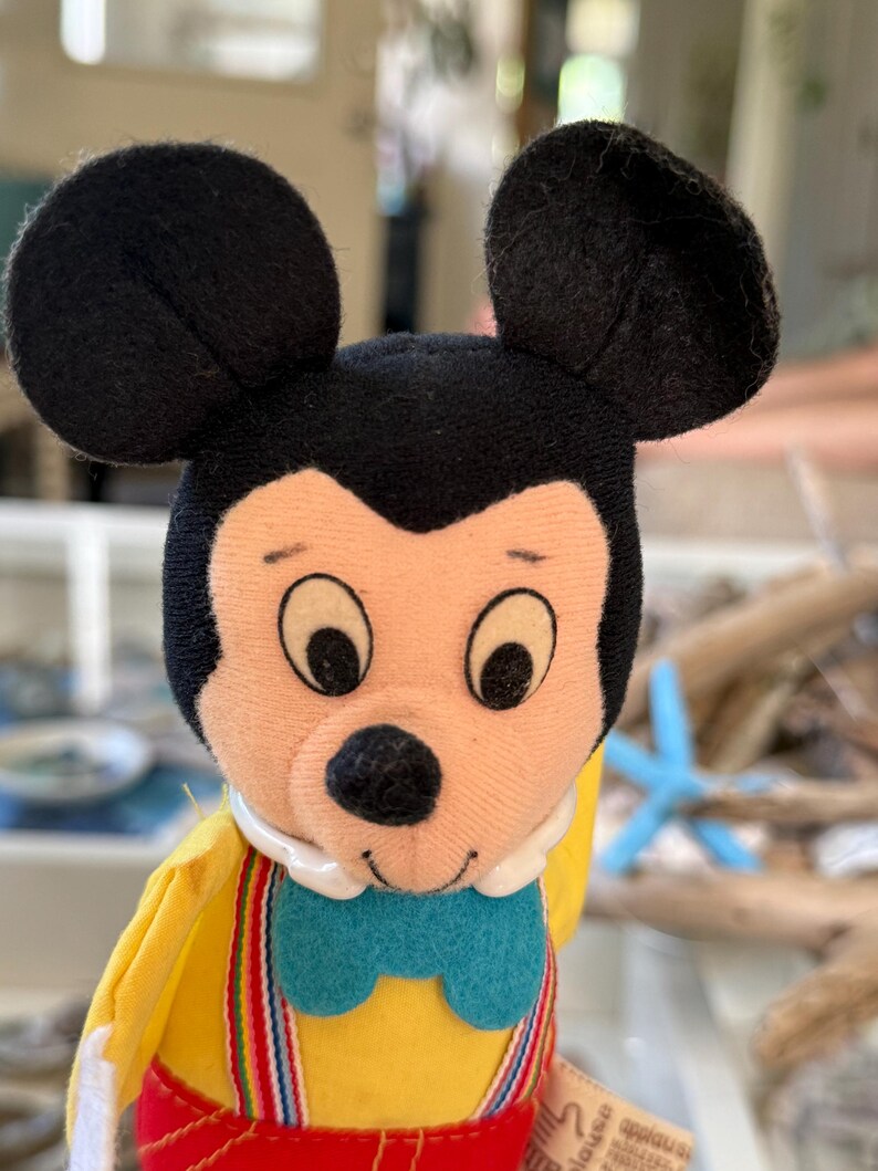 May include: A plush Mickey Mouse doll featuring large black ears, a peach-colored face, and a black nose. The doll wears a yellow shirt, a bright blue bowtie, and red pants held up by multicolored striped suspenders with a white collar.