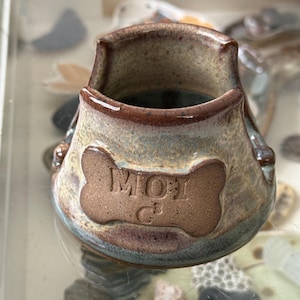 May include: A ceramic pet bowl with a bone-shaped design and the word "MOT" embossed on the front. The bowl has a brown rim and a blue and brown exterior. The bowl is designed for pets.