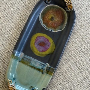 May include: A handmade ceramic serving platter with a dark blue-gray base. It features two circular designs with yellow, red, and purple hues. The platter has a light blue-green section and a decorative handle with a textured design.