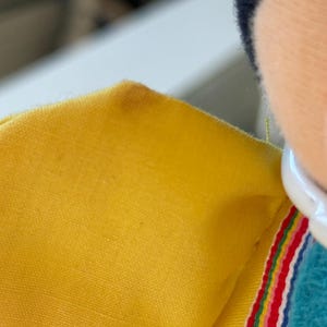 May include: A yellow fabric garment featuring a white collar and a decorative trim with horizontal stripes in red, yellow, green, and blue. The fabric has a visible woven texture, and a small dark spot is present on the upper yellow section.
