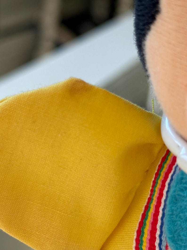 May include: A yellow fabric garment featuring a white collar and a decorative trim with horizontal stripes in red, yellow, green, and blue. The fabric has a visible woven texture, and a small dark spot is present on the upper yellow section.