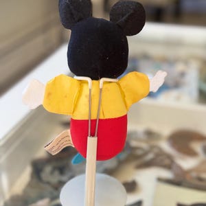 May include: A plush Mickey Mouse character figure viewed from the back, featuring a black head with round ears, a yellow shirt, and red pants. The figure is supported by a white metal display stand with two thin prongs holding the torso.